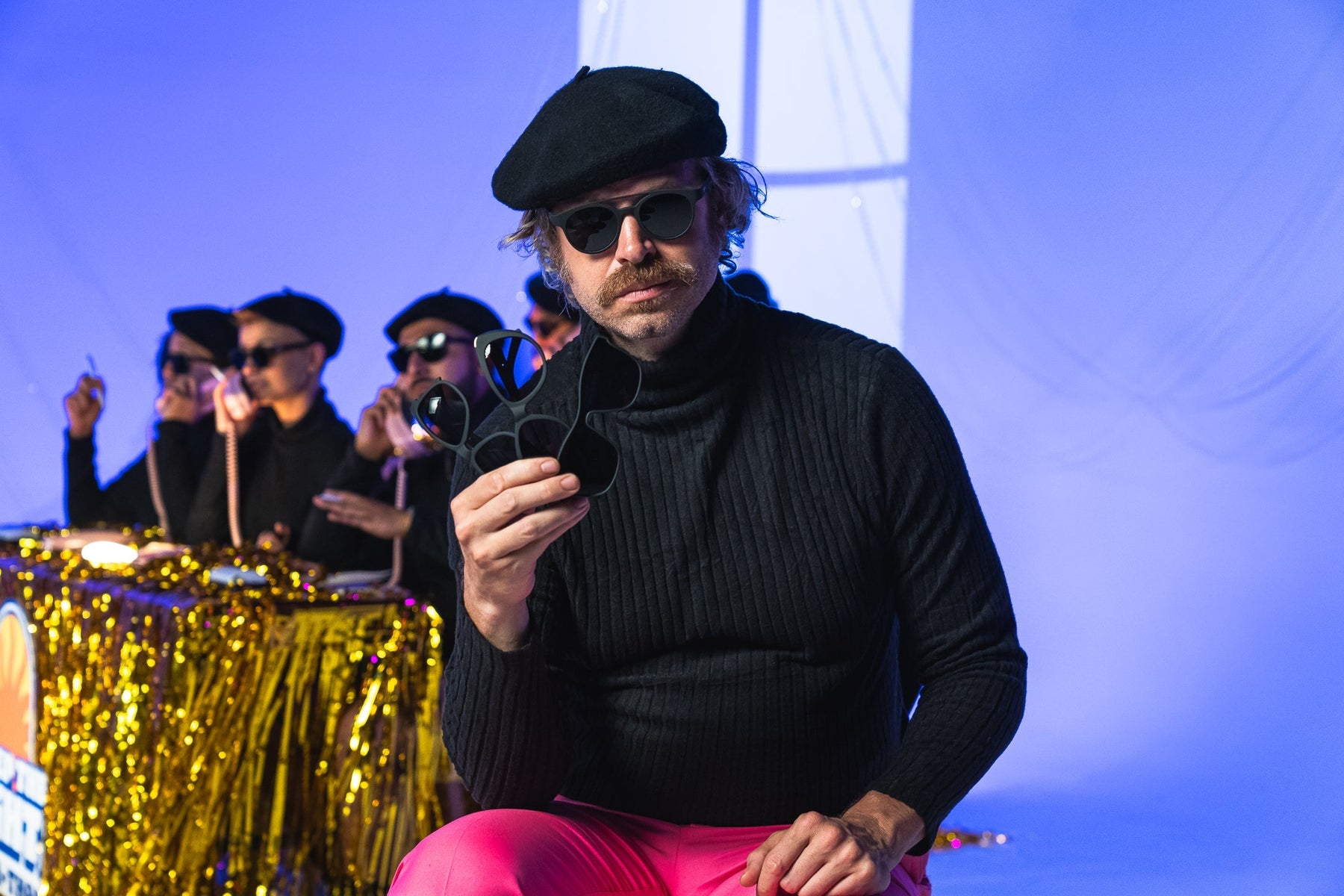 Person in black sweater and a beret holding goodr sunglasses on stage.