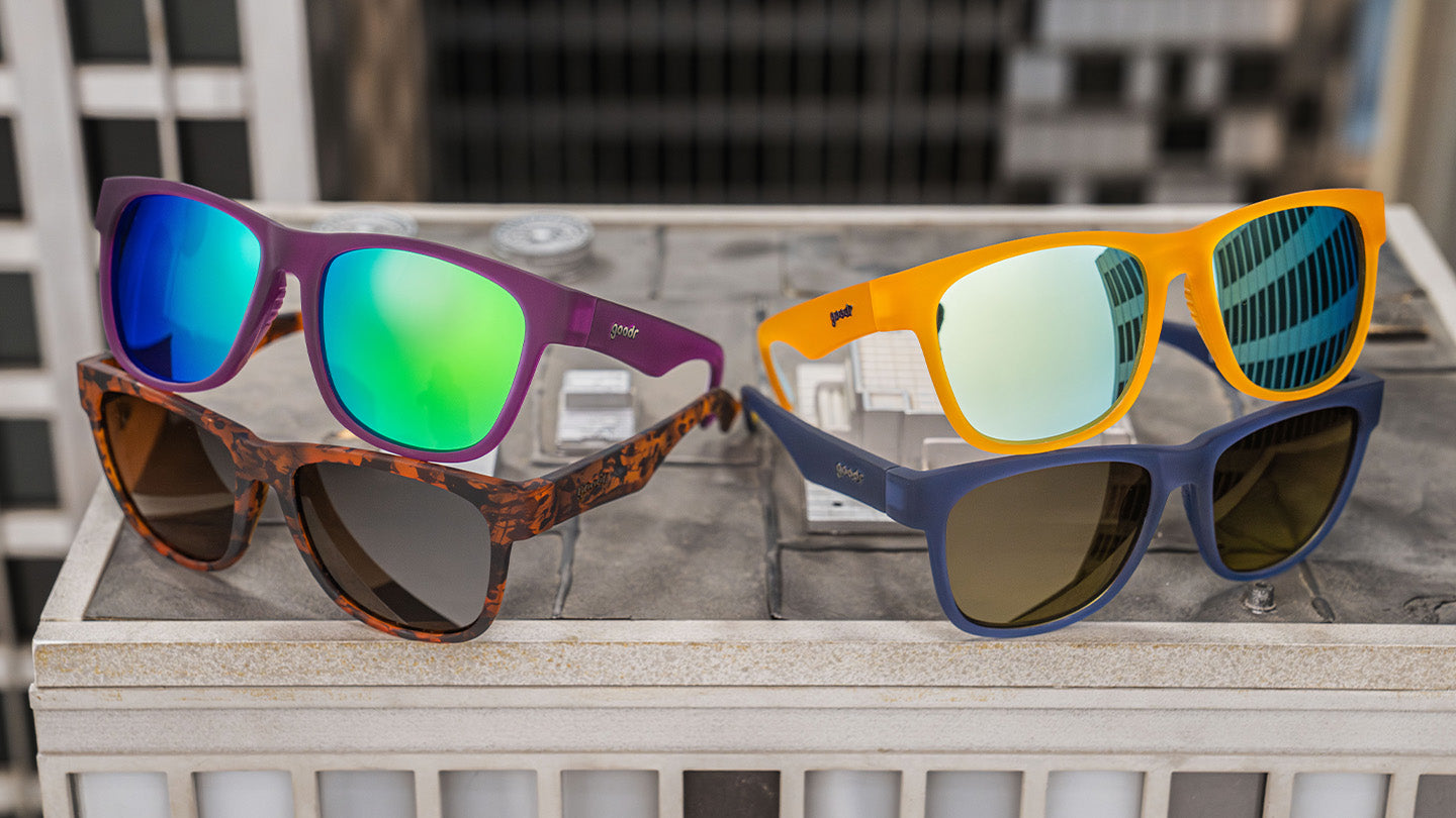 Four pairs of sunglasses displayed on a rooftop surface, featuring purple, tortoiseshell, orange, and blue frames, each with reflective or tinted lenses.