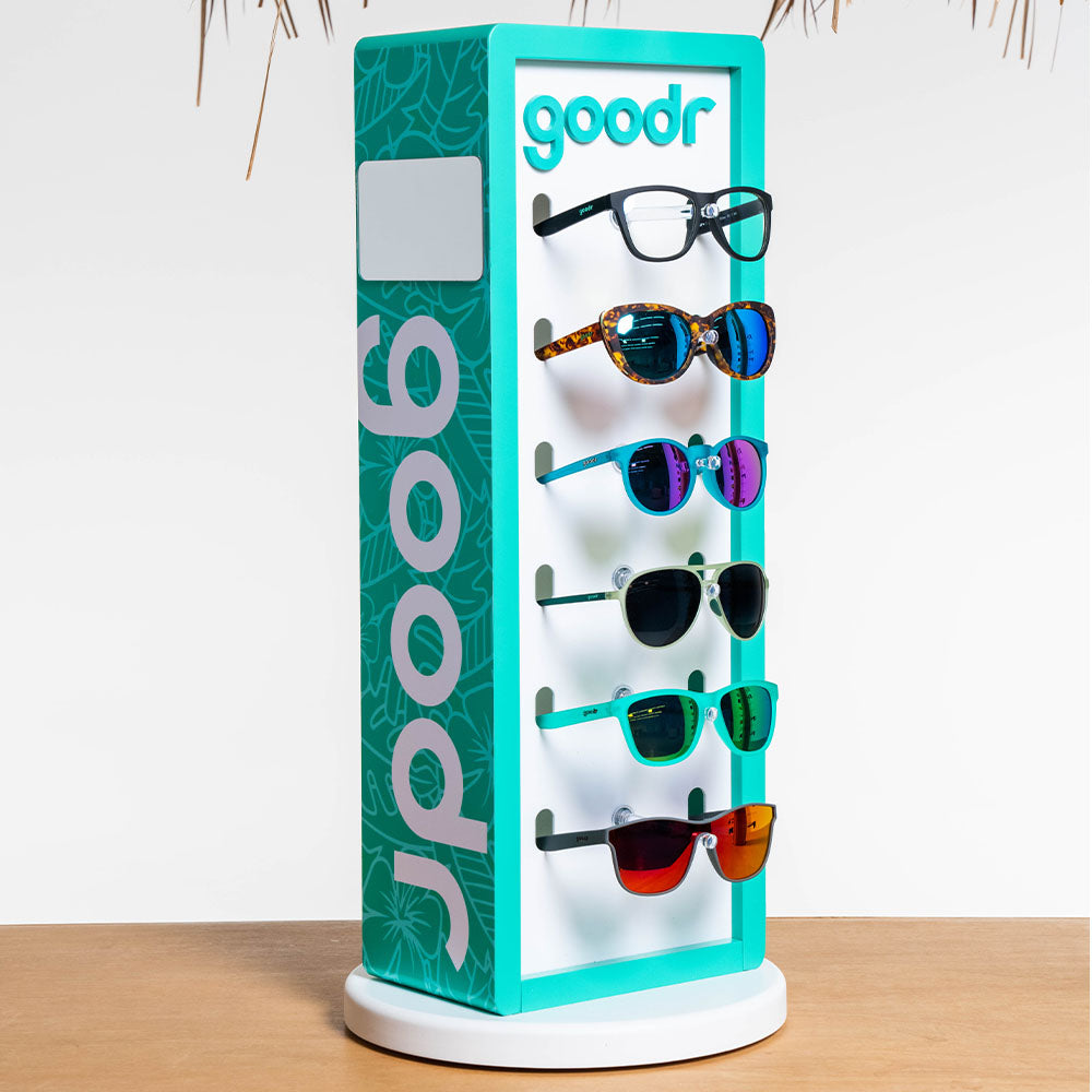 A teal and white "goodr" branded display stand showcasing six pairs of sunglasses and eyeglasses. The stand has various styles, including colorful and reflective lenses, positioned neatly on pegs against a white background.
