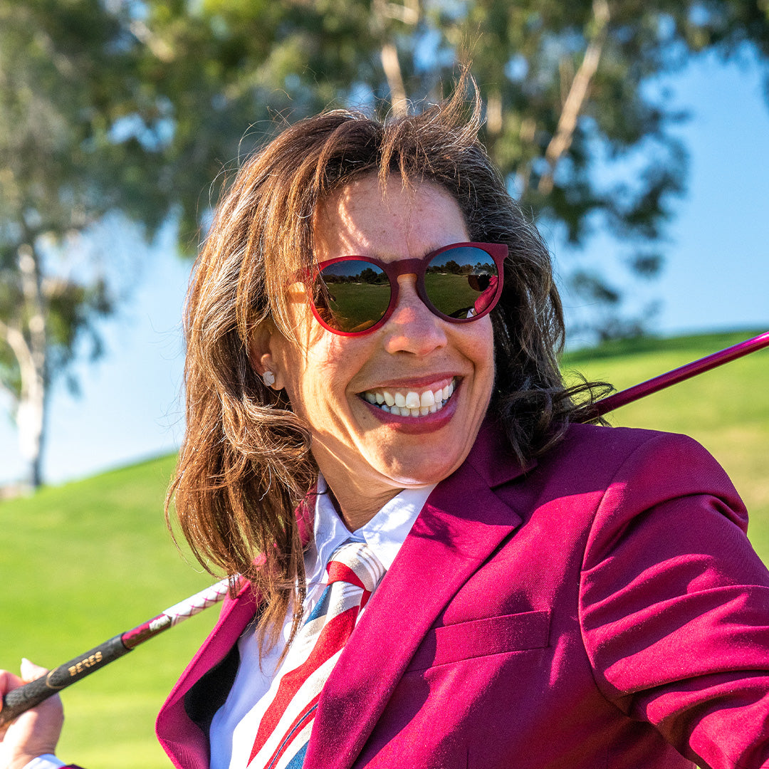 A woman dressed in a burgundy blazer and tie is smiling while holding a golf club over her shoulder. She is wearing red-framed sunglasses with reflective lenses that capture the golf course in front of her.