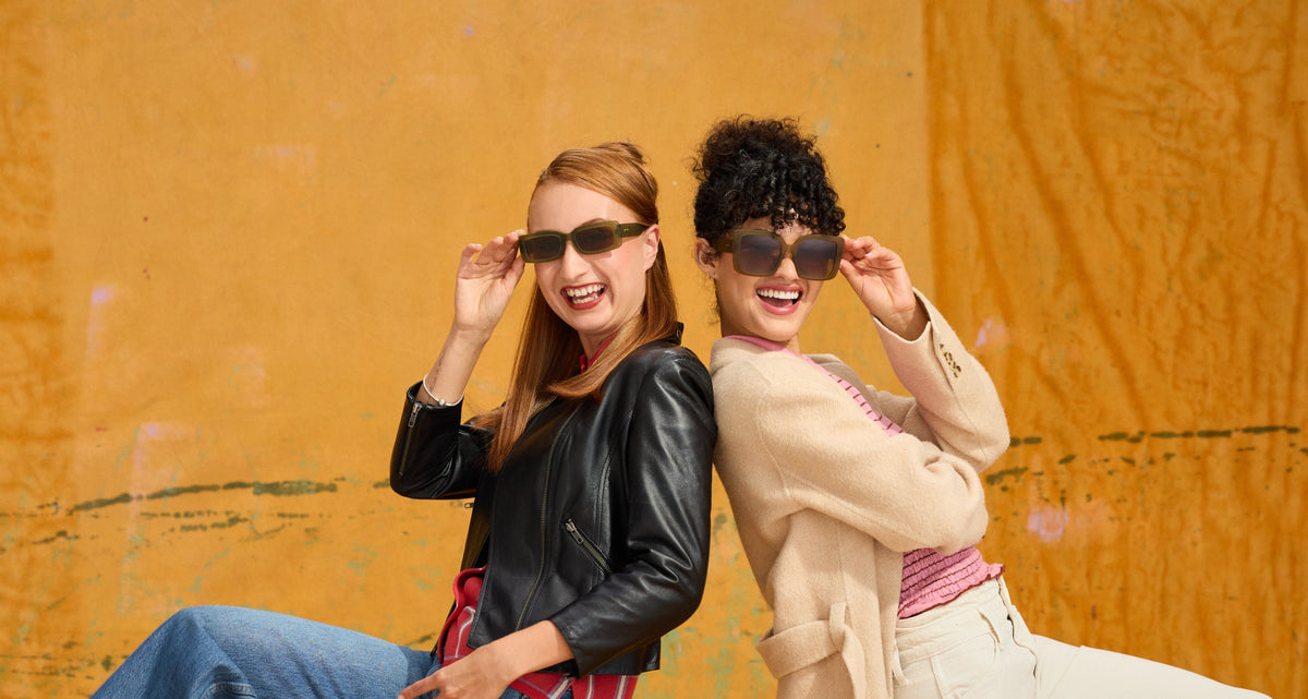Two women posing back-to-back in stylish goodr sunglasses from the Fall Winter 2026 Collection, a black leather jacket, blue jeans, and a beige trench coat against a textured orange wall.