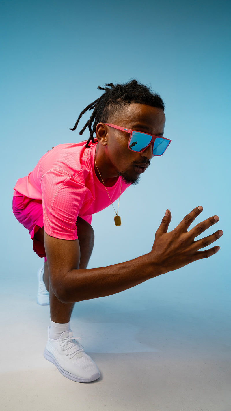 A man in a pink outfit, posed to begin running, wearing pink framed, square shaped sunglasses.