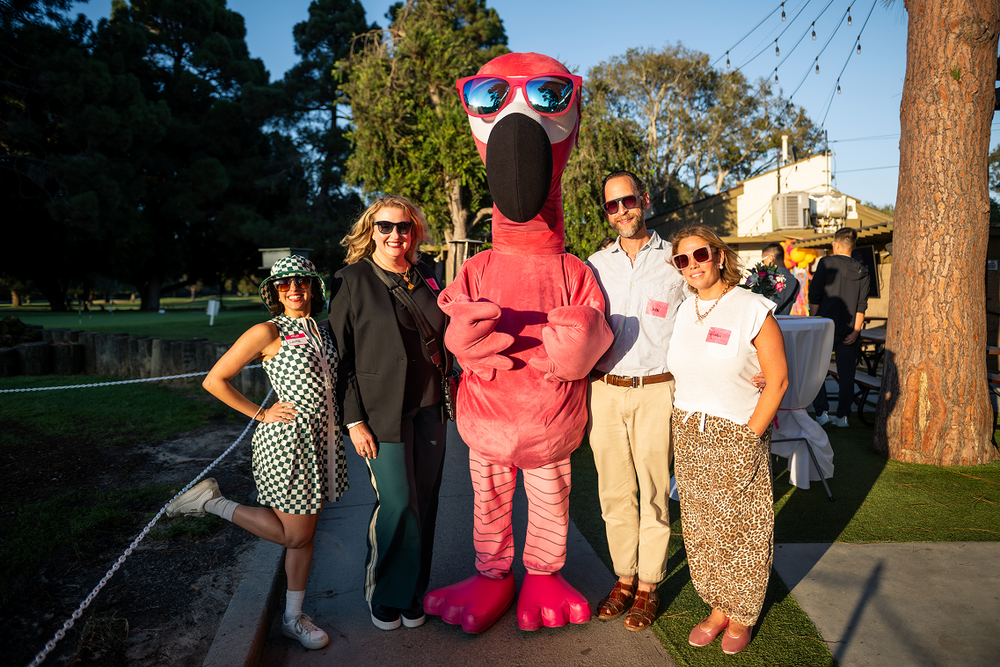 Group of people posing with a pink flamingo mascot in an outdoor setting.