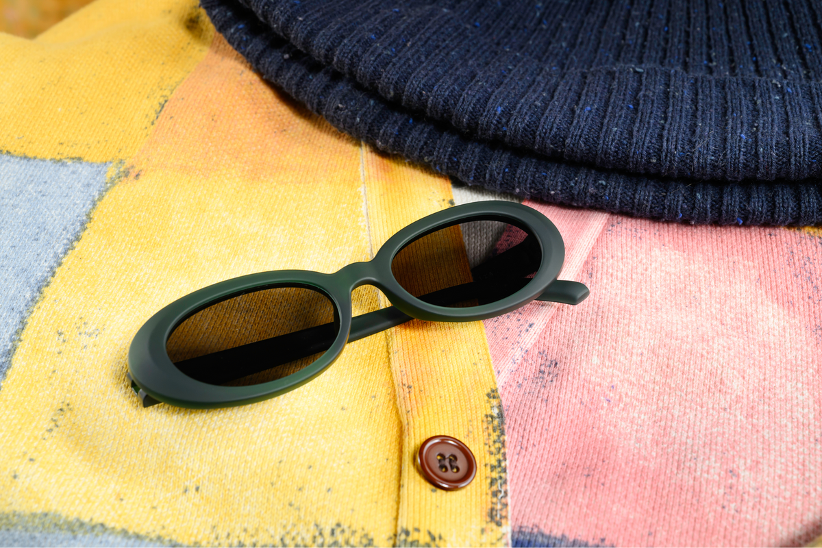 Beat G oval sunglasses with a matte dark green frame and dark lenses, resting on a colorful patchwork knit cardigan with a navy blue ribbed beanie partially visible at the top.