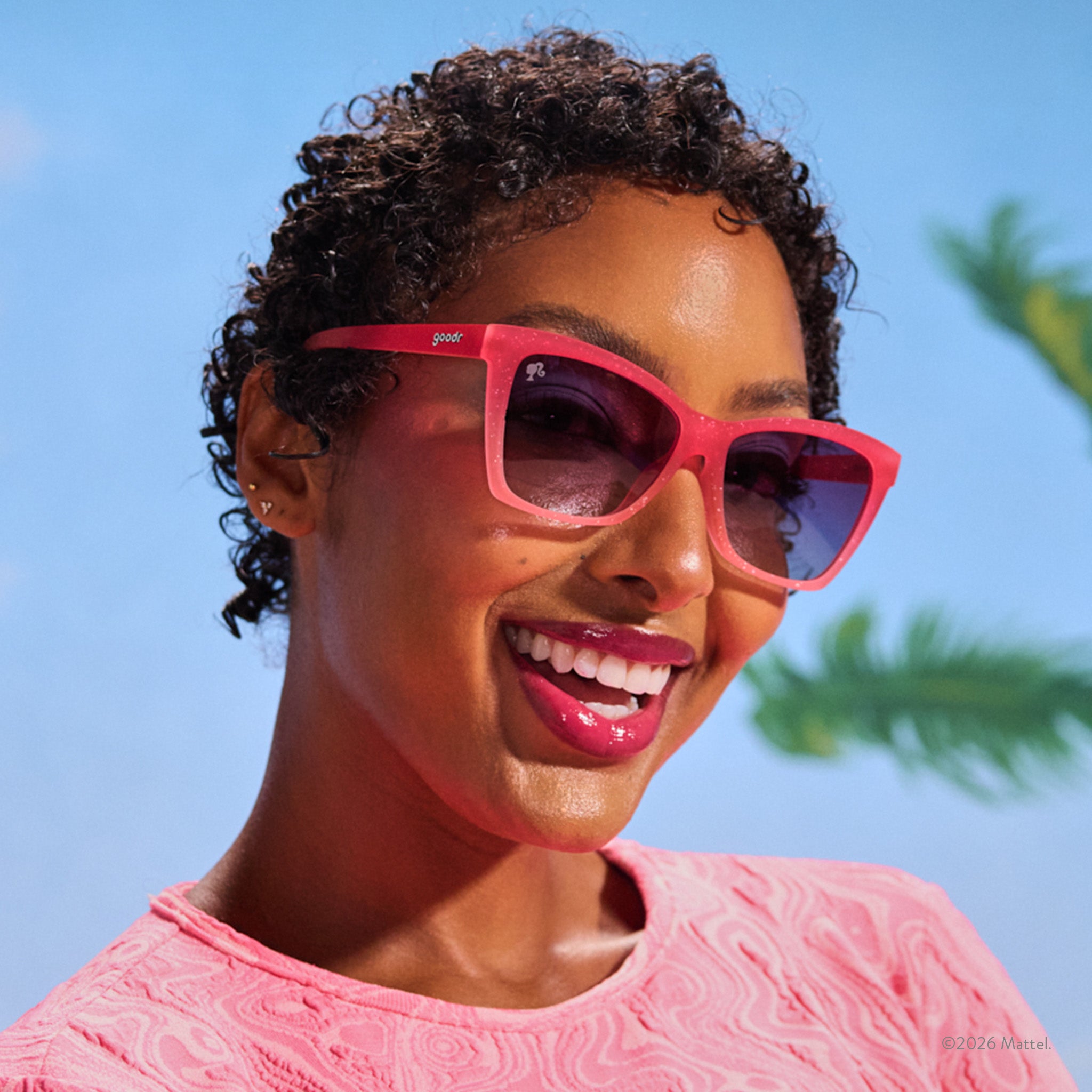 A young woman with short, curly dark hair smiles confidently in glittery pink, angular cat-eye Barbie™ sunglasses.