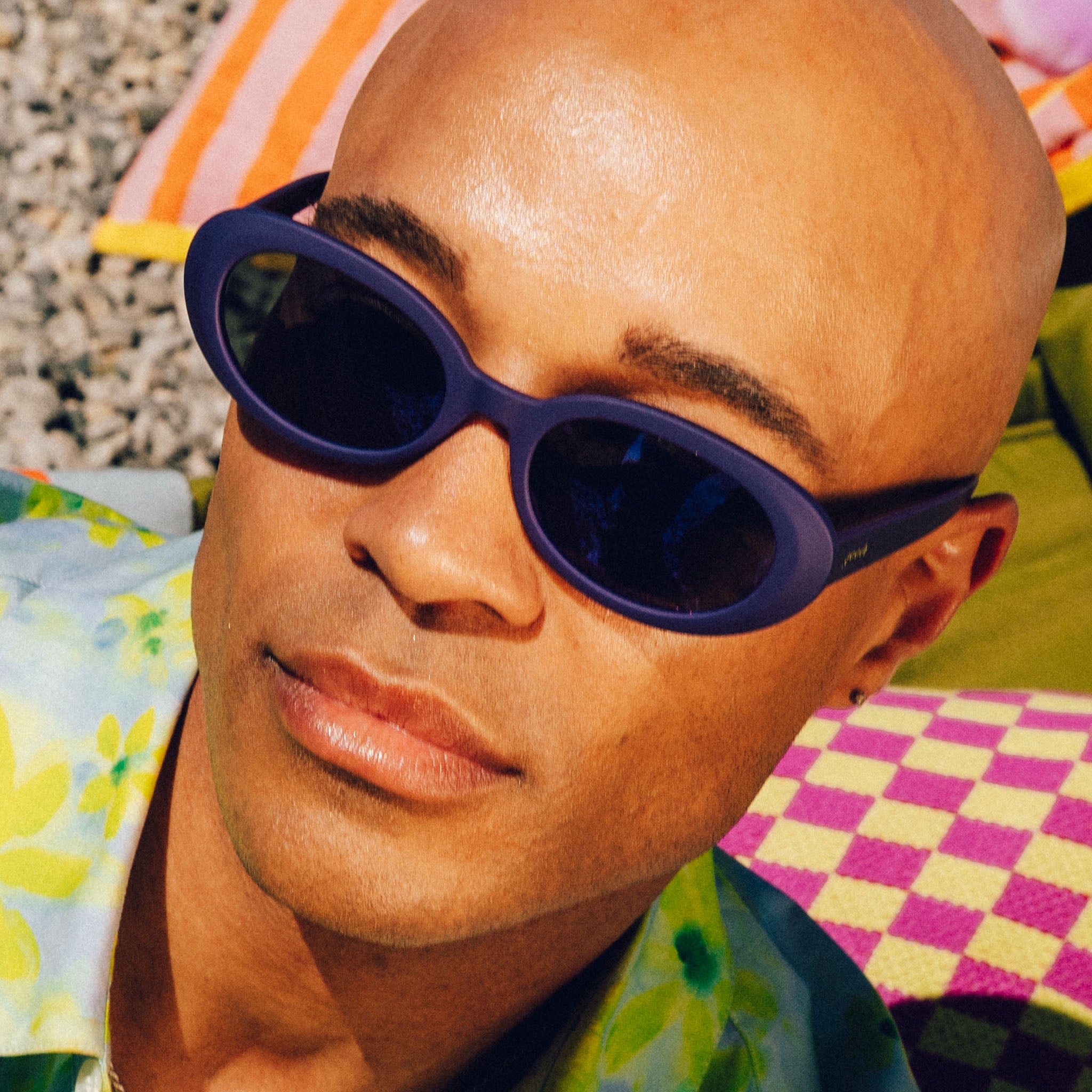 A bald man in a floral shirt wears deep purple, oval-shaped sunglasses with reflective purple lenses. Wild patterns play in the background.