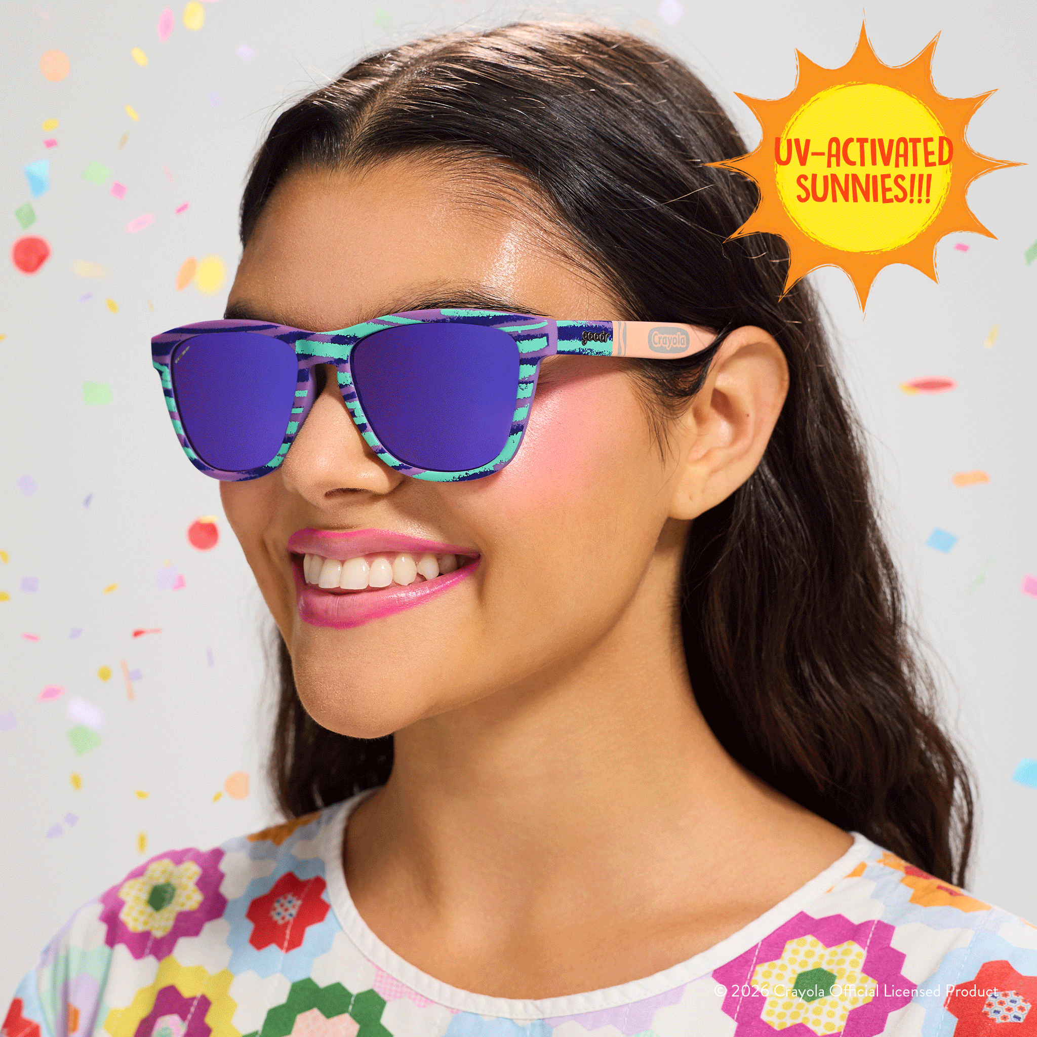 Young woman smiles, wearing Crayola goodr UV-activated sunglasses with green & purple frames & purple reflective lenses.