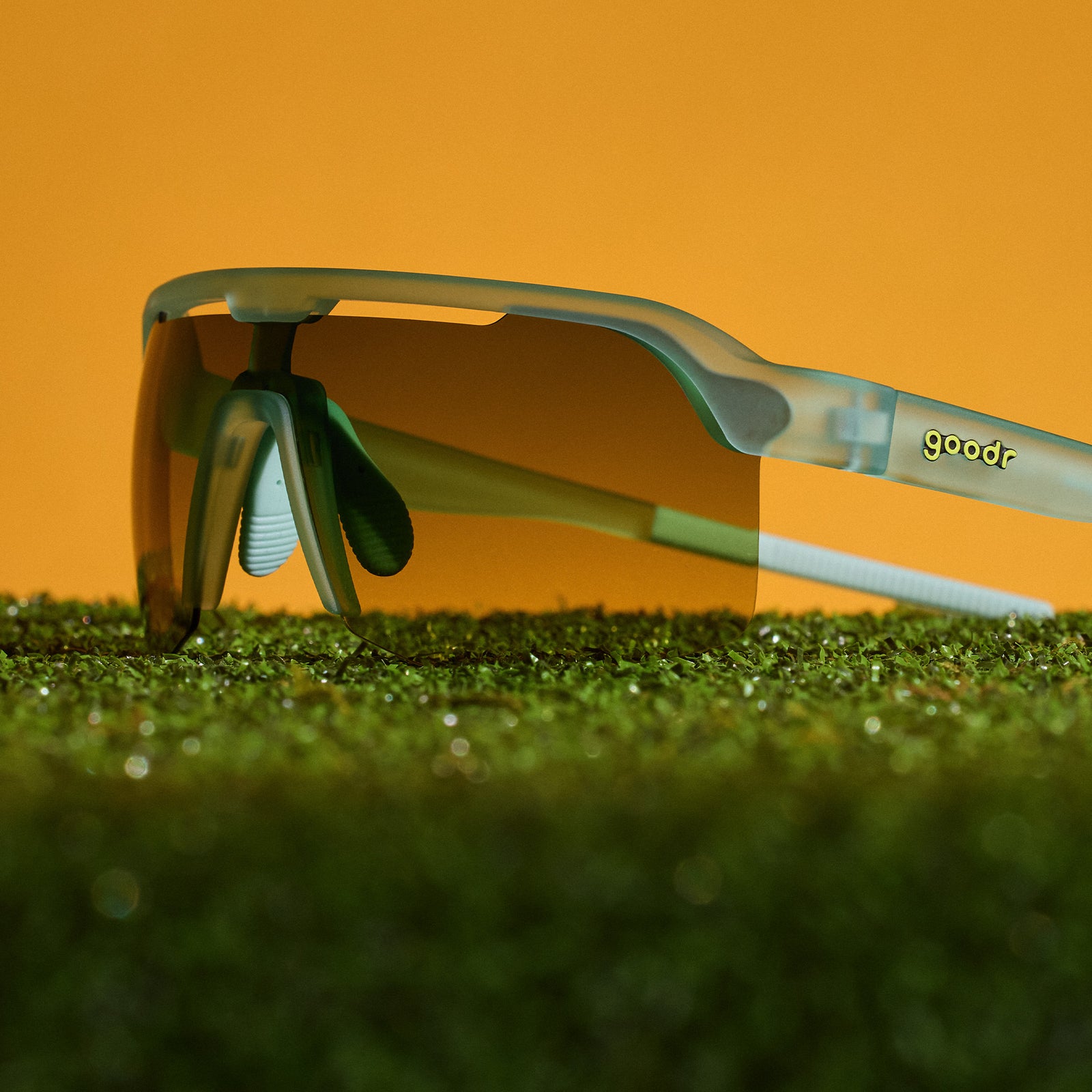 Goodr sport sunglasses with a translucent light green frame and orange-tinted lenses, resting on green grass against a solid orange background.