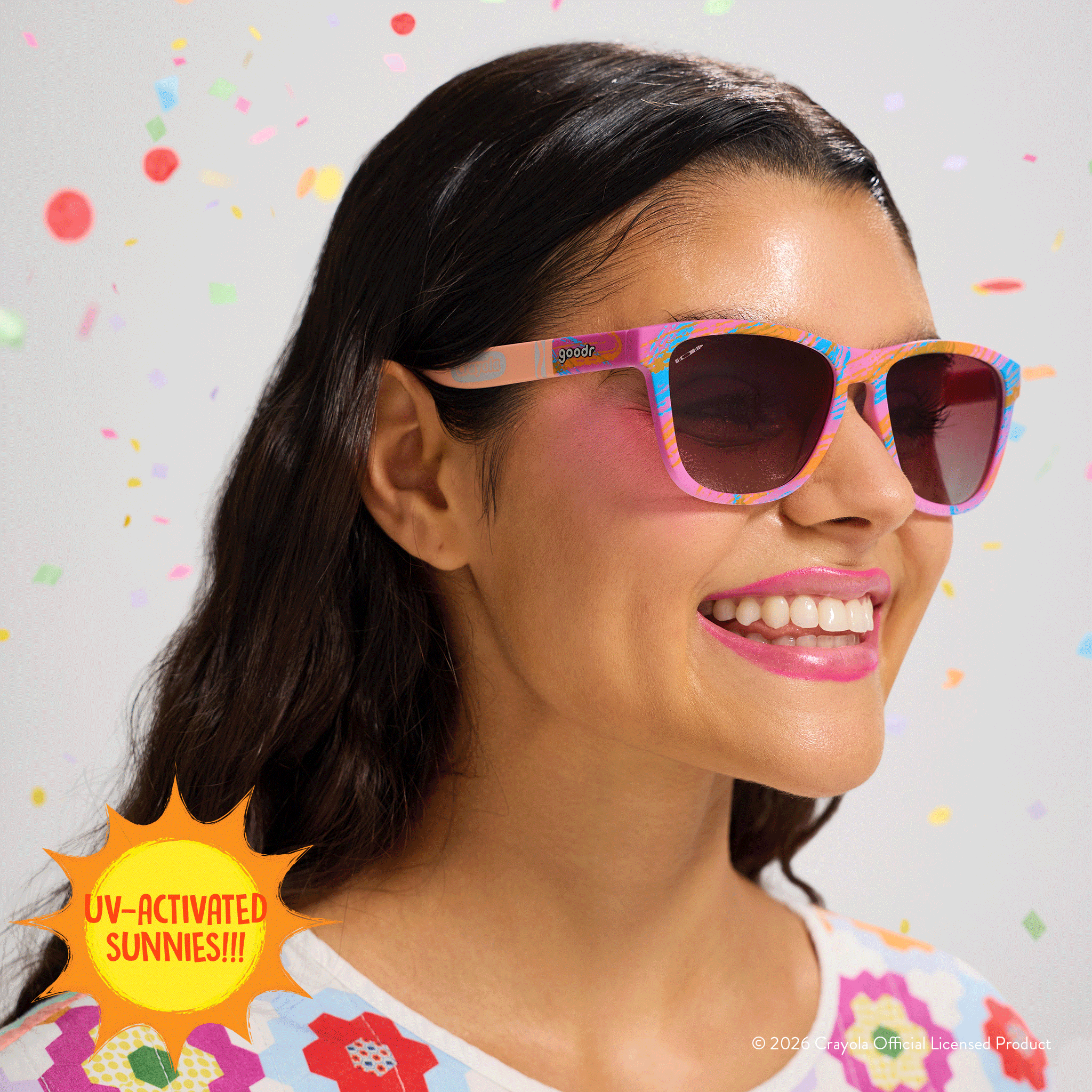Young woman smiles, wearing Crayola goodr sunglasses with tan-to-magenta frames and rose ocean gradient lenses.