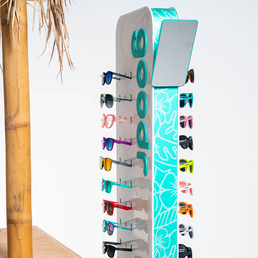 Freestanding goodr sunglasses display rack showcasing various colorful sunglasses on both sides. The display features a teal floral pattern and branding on the sides, with tropical decor including a bamboo pole partially visible on the left.