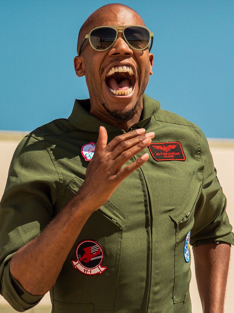 Man wearing aviator sunglasses and a green flight suit, laughing enthusiastically under a bright blue sky, with various patches on his suit adding to the military-inspired look.