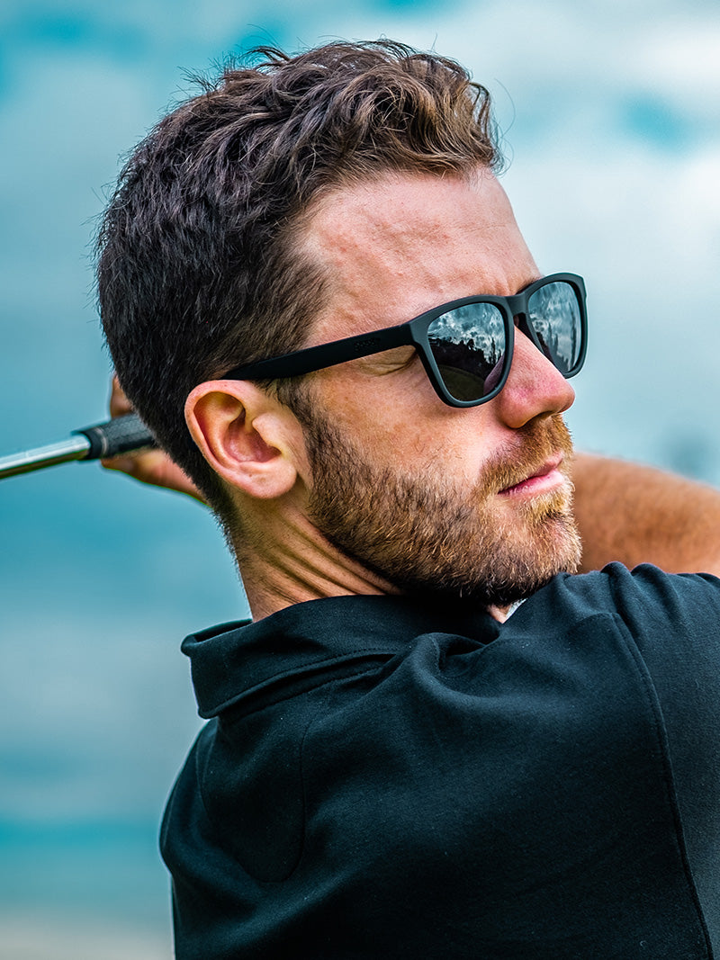 A man golfing, wearing a black polo shirt and black square sunglasses.