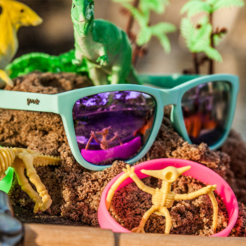 goodr sunglasses with purple lenses and teal frames, sitting on sand surrounded by dinosaur toys