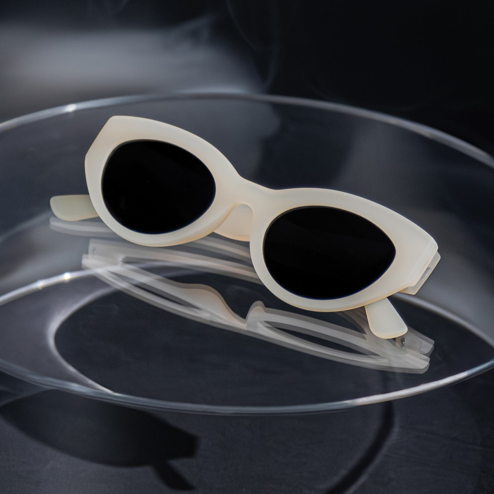Close-up of translucent soft cream, retro-inspired angled cat-eye sunglasses with black non-reflective lenses, folded on a clear table.