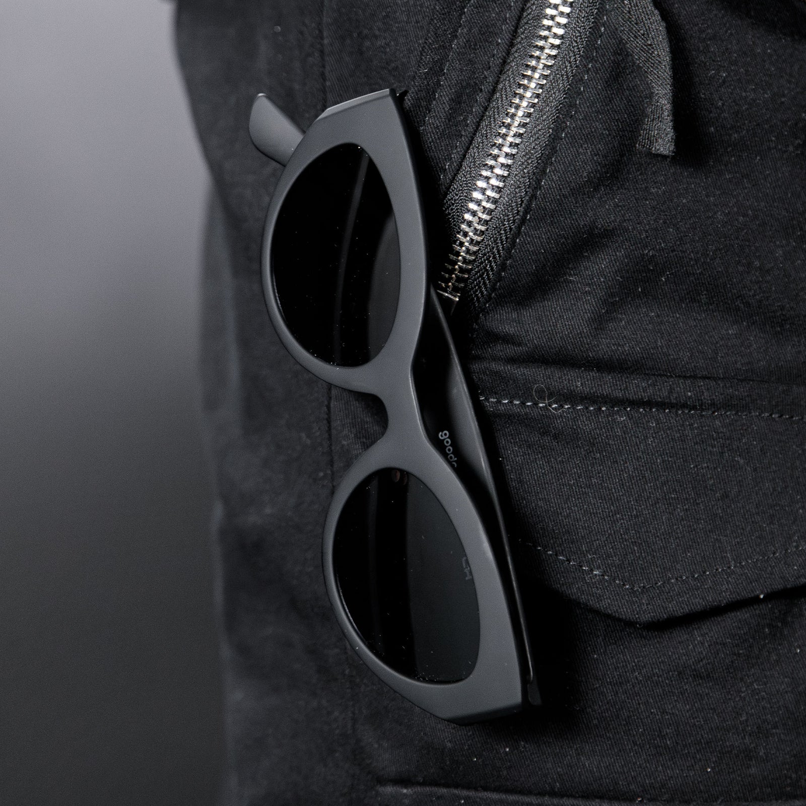 Close-up of translucent black, retro-inspired angled cat-eye sunglasses with non-reflective lenses hanging from a black zippered pant pocket.