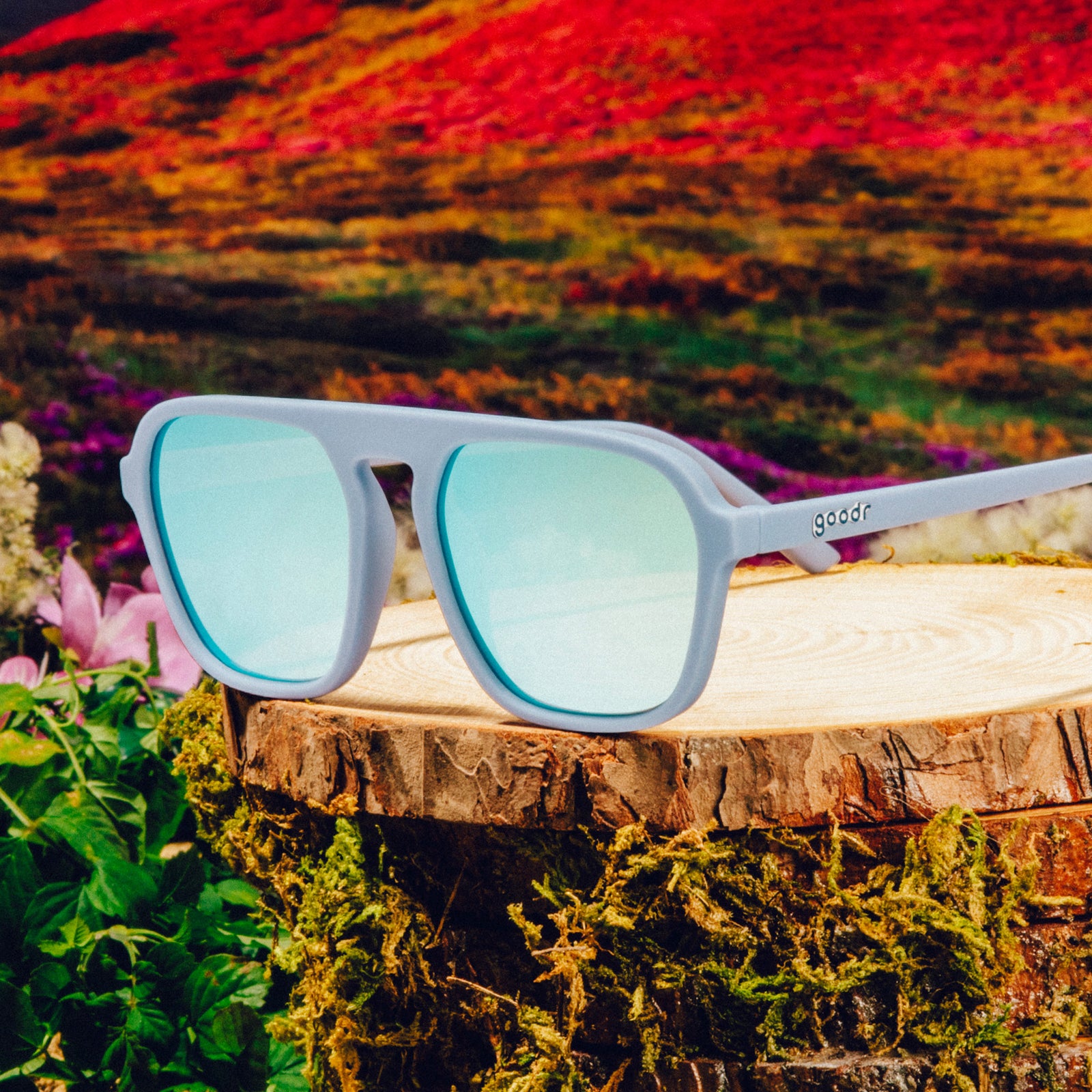 Three-quarter angle view of light blue retro-inspired aviator sunglasses with blue reflective lenses on a tree trunk against a meadow backdrop.