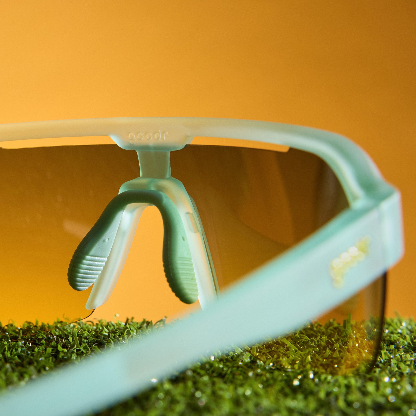 Close-up of sport sunglasses with a translucent mint green frame and tinted lens, resting on green grass against a warm orange background.