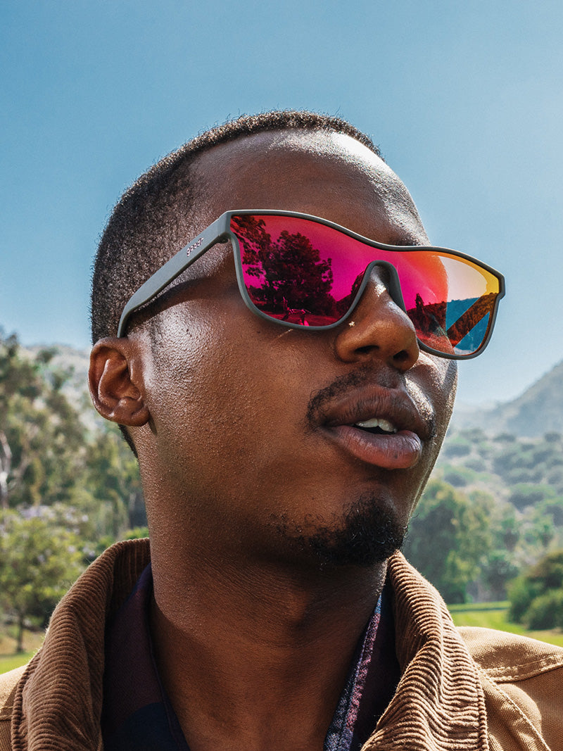 A man wearing a brown jacket, wearing black, single lens sunglasses with reflective, red to yellow ombre lenses.