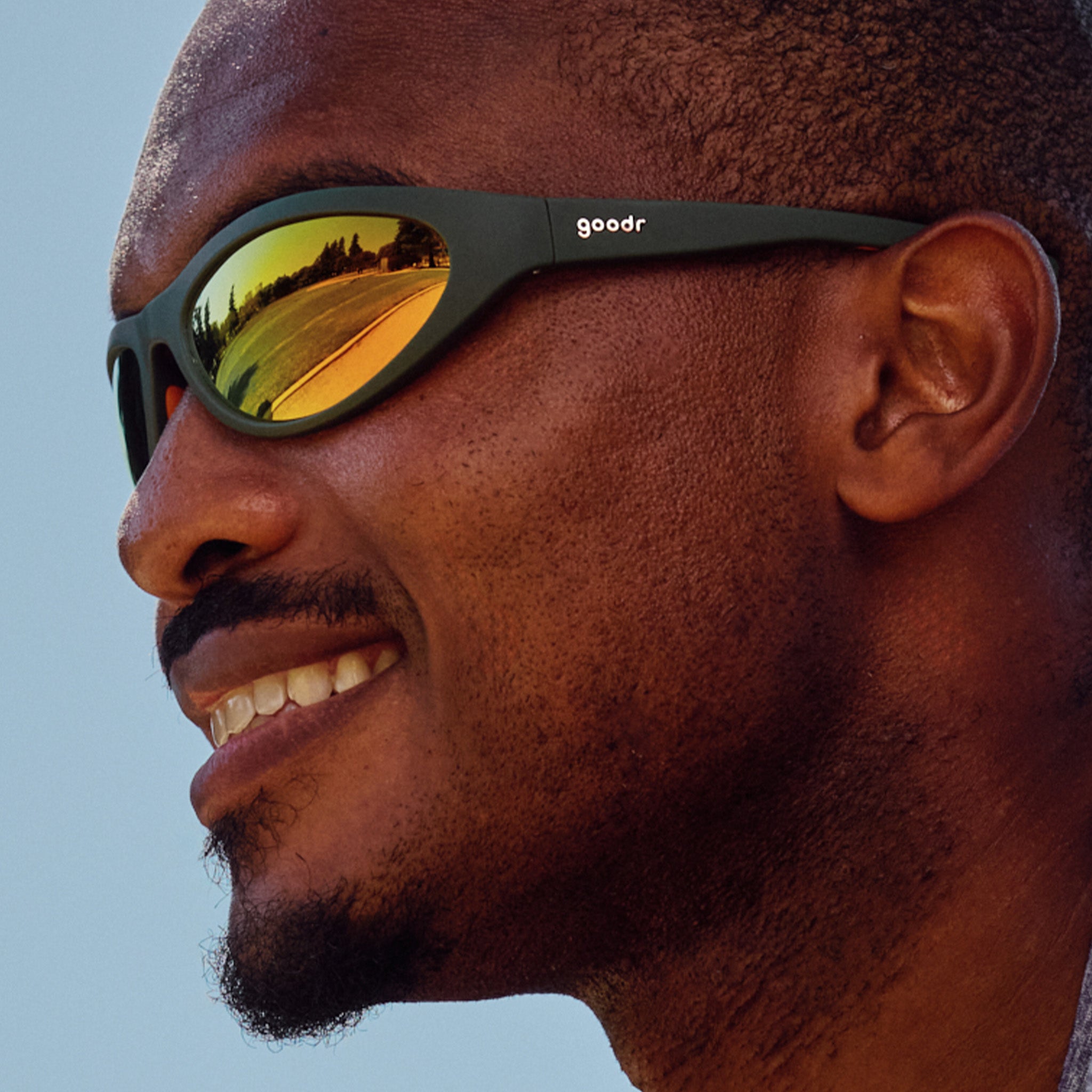 Close-up of a smiling person wearing green bug eyed sunglasses with reflective yellow lenses, outdoors on a sunny day.