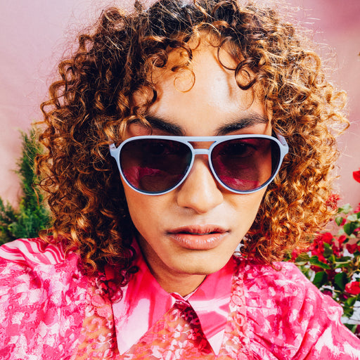 Front view of woman wearing light blue aviator sunglasses with purple gradient lenses against a meadow backdrop.