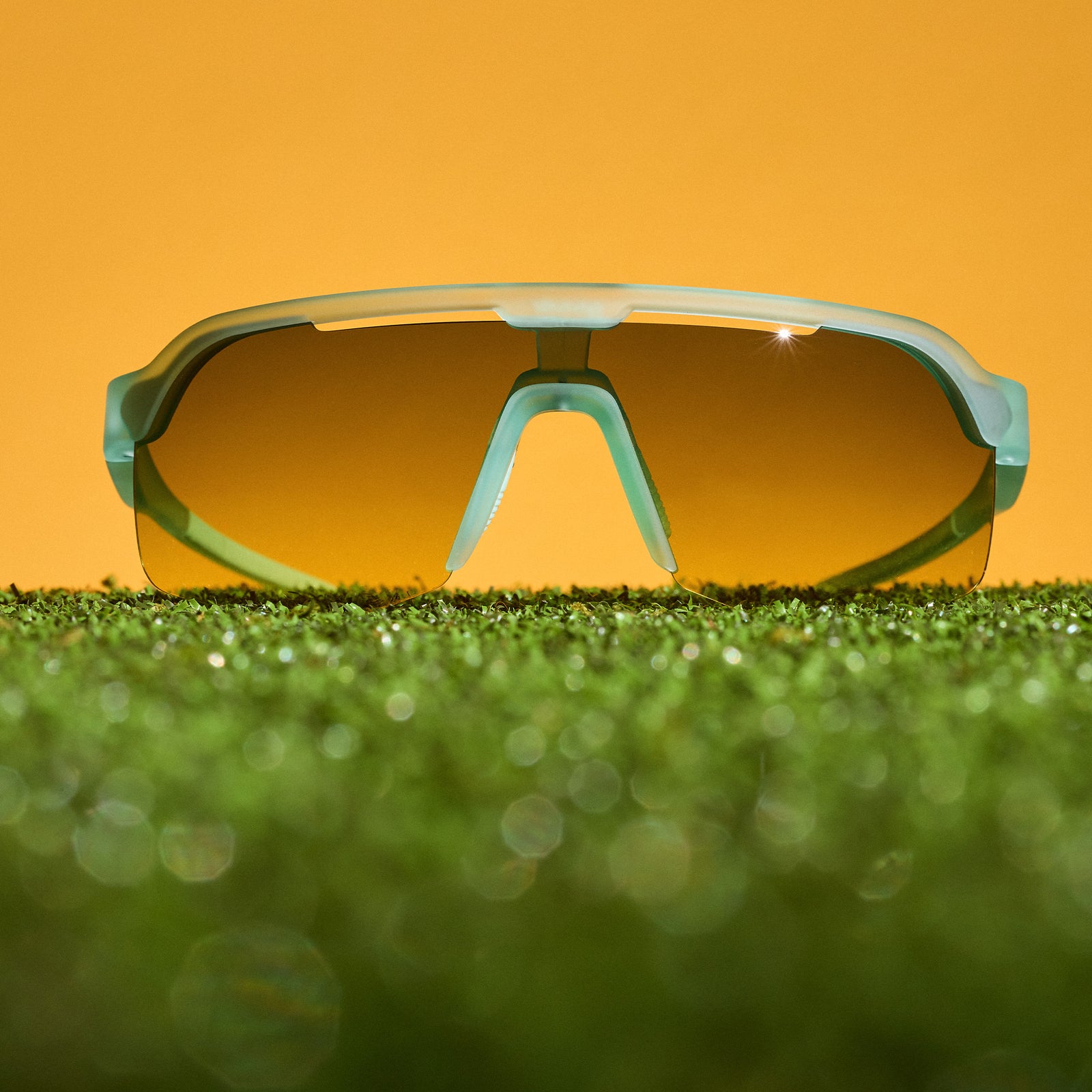 Sport sunglasses with a light blue frame and a yellow-orange tinted lens, resting on green grass against a solid orange background.