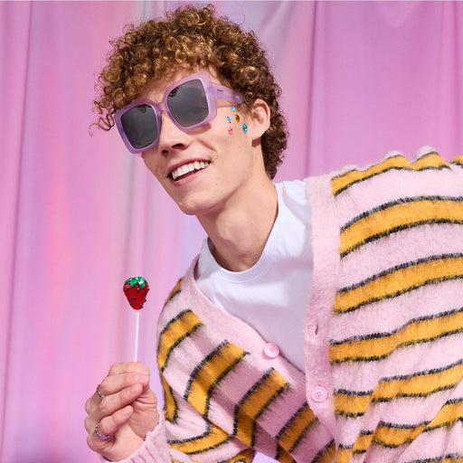 A young man with curly hair smiles while holding a lollipop that looks like a strawberry. He wears pale purple Amp G sunnies.