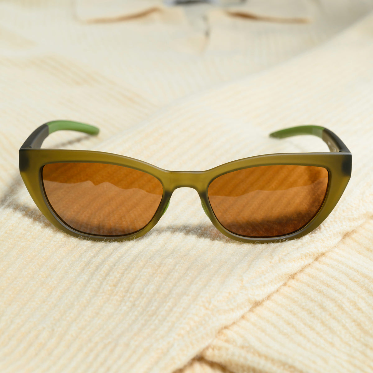A pair of translucent olive-green cat-eye sunglasses with brown non-reflective lenses sits on a cream-colored sweater.