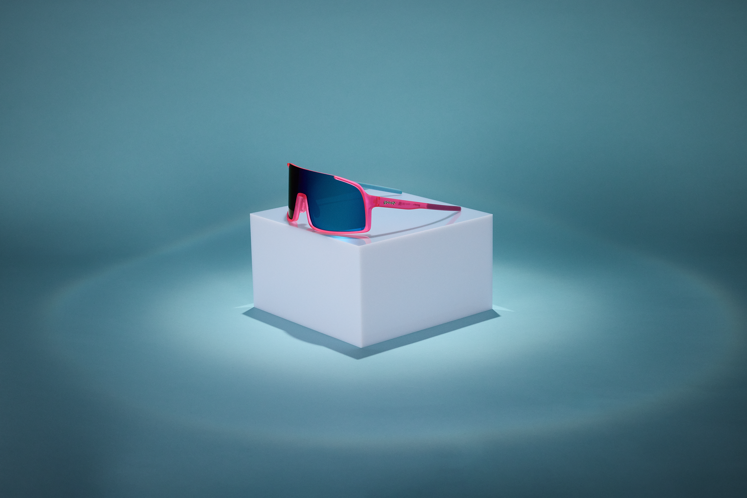 A pair of wraparound goodr sunglasses with pink frames and blue lenses, perched on top of a white cube on a blue background.