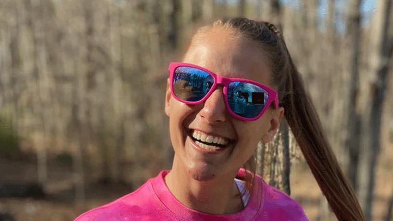 A woman with a pony tail smiling, wearing a bright pink shirt and sunglasses with bright pink frames.