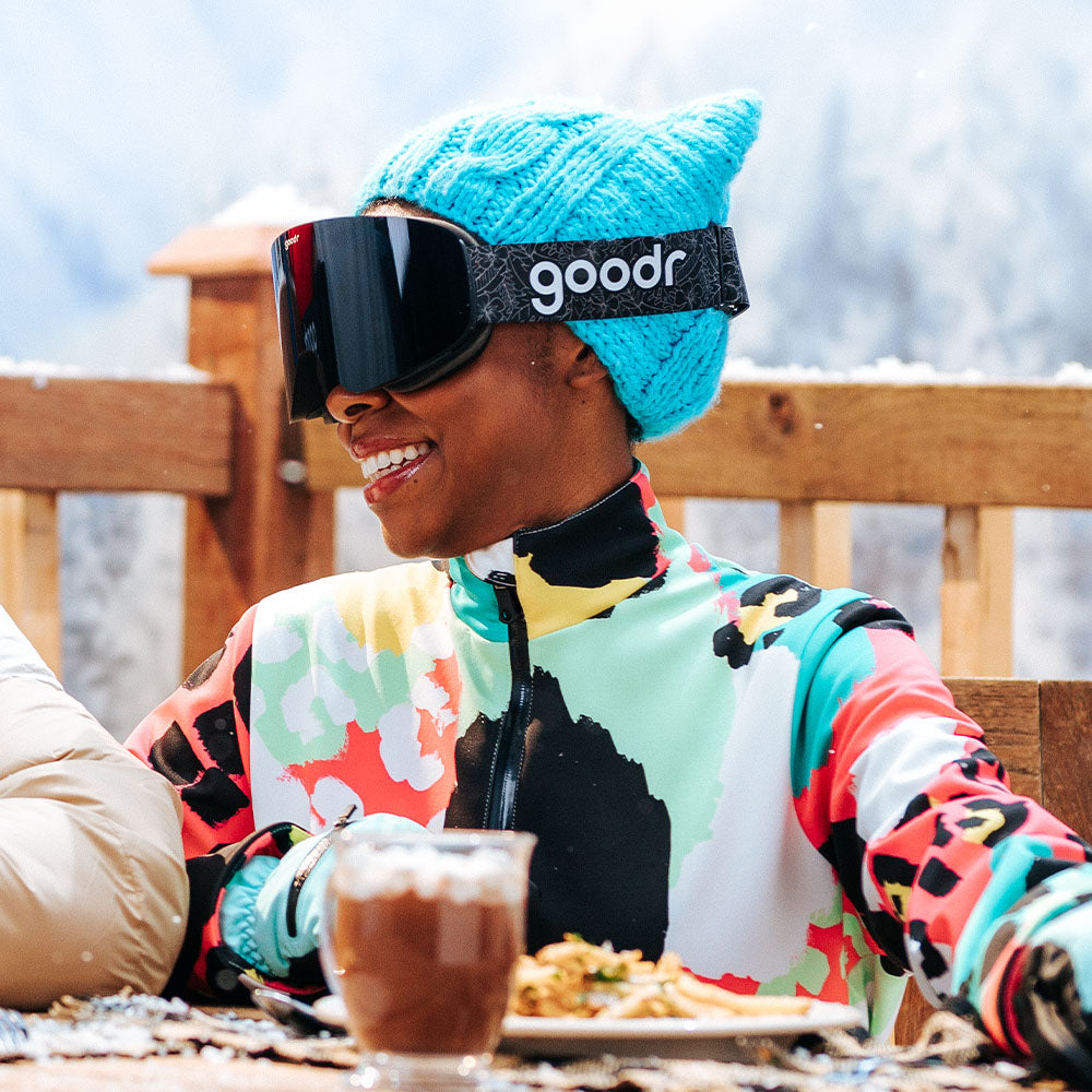 A woman wearing black goodr snowboarding goggles and a blue beanie, sitting outside eating food