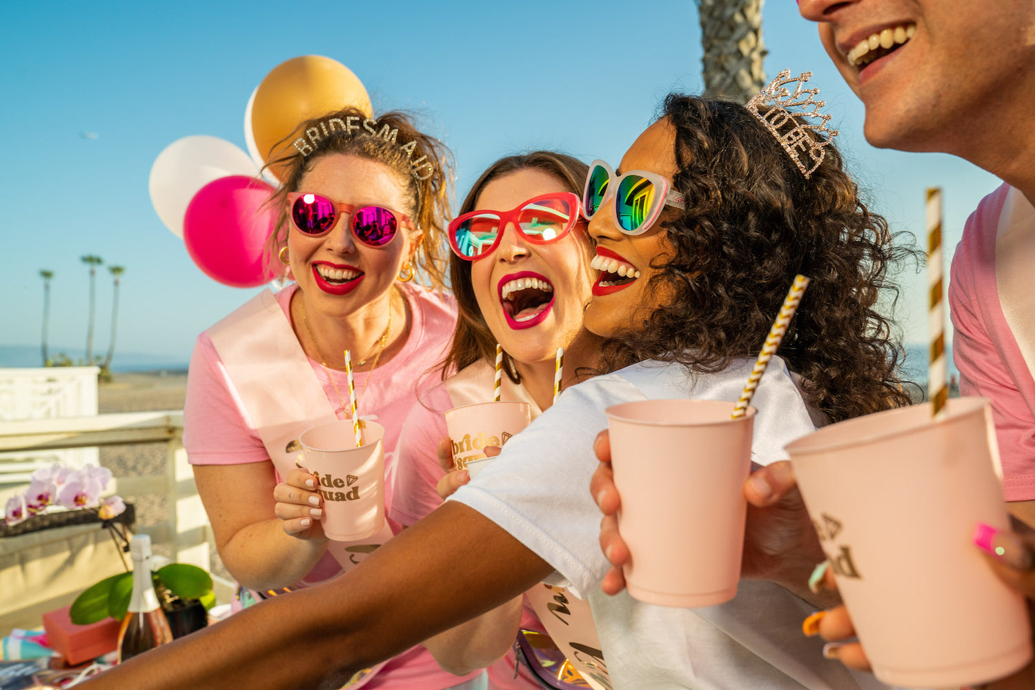 3 women wearing different colored sunglasses, smiling and laughing, holding matching pink and gold cups that read "Bride Squad".