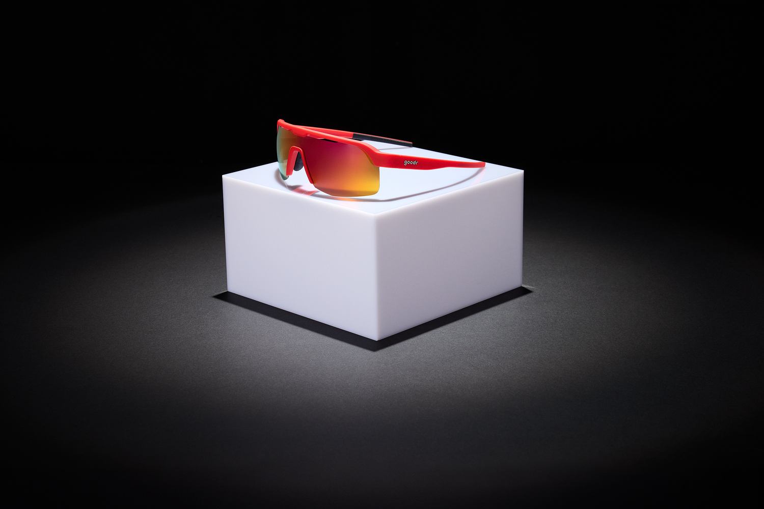 Red sports sunglasses with reflective orange lenses, displayed on an illuminated white pedestal against a dark background for a dramatic effect.