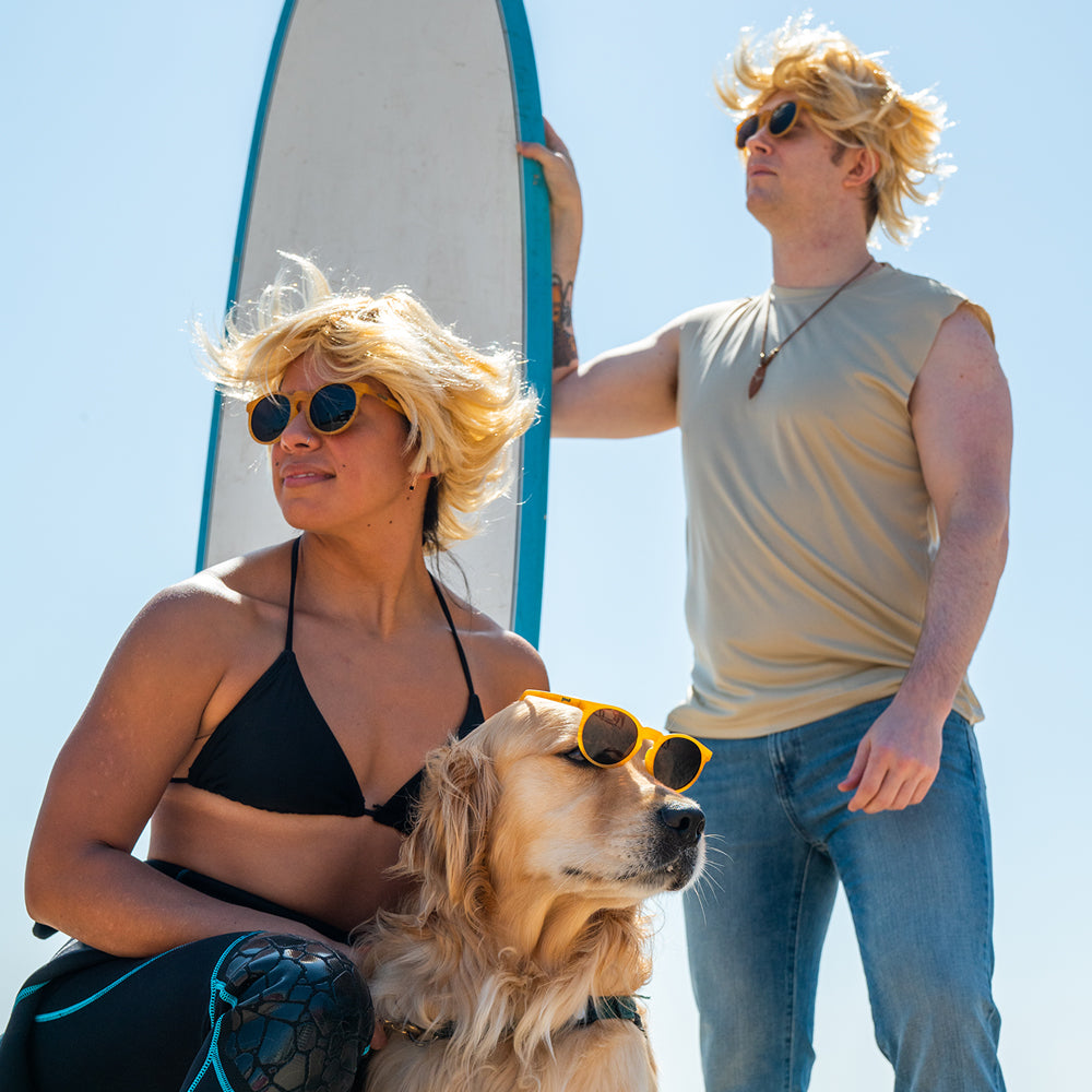 A woman, man, and a golden retriever all wearing blonde wigs and sunglasses, posing by a surfboard on a sunny beach.