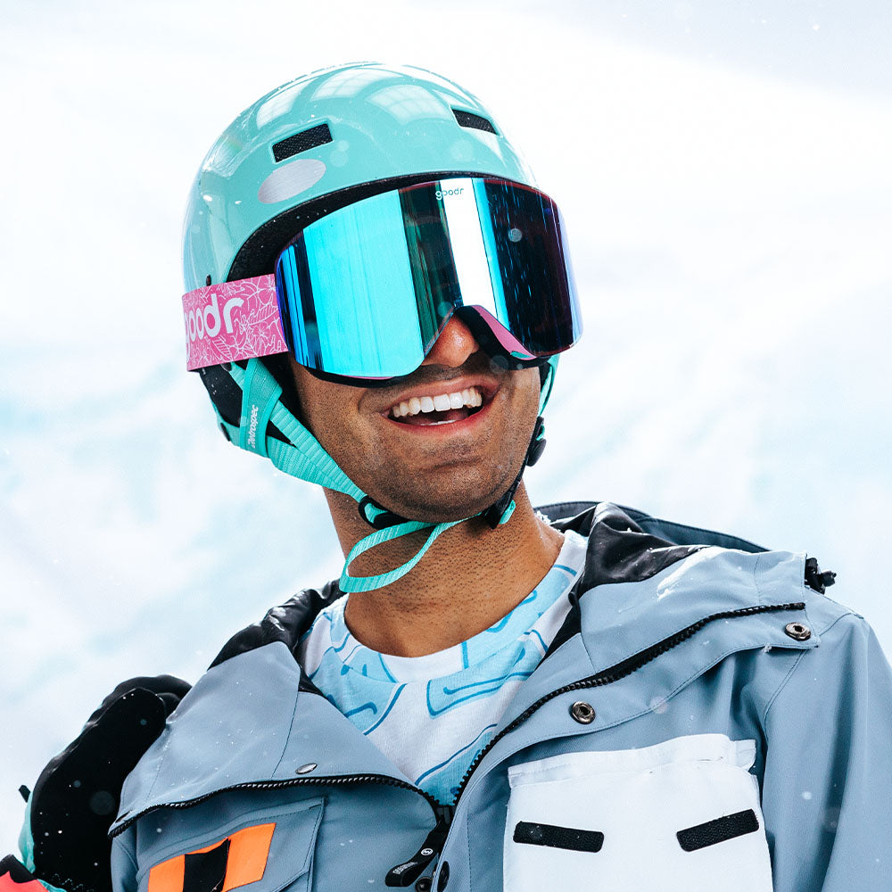 A man wearing a light blue helmet, reflective blue ski goggles, and a gray ski jacket smiles while standing in the snow. His goggles have a pink "goodr" branded strap, and the background shows a bright snowy landscape.