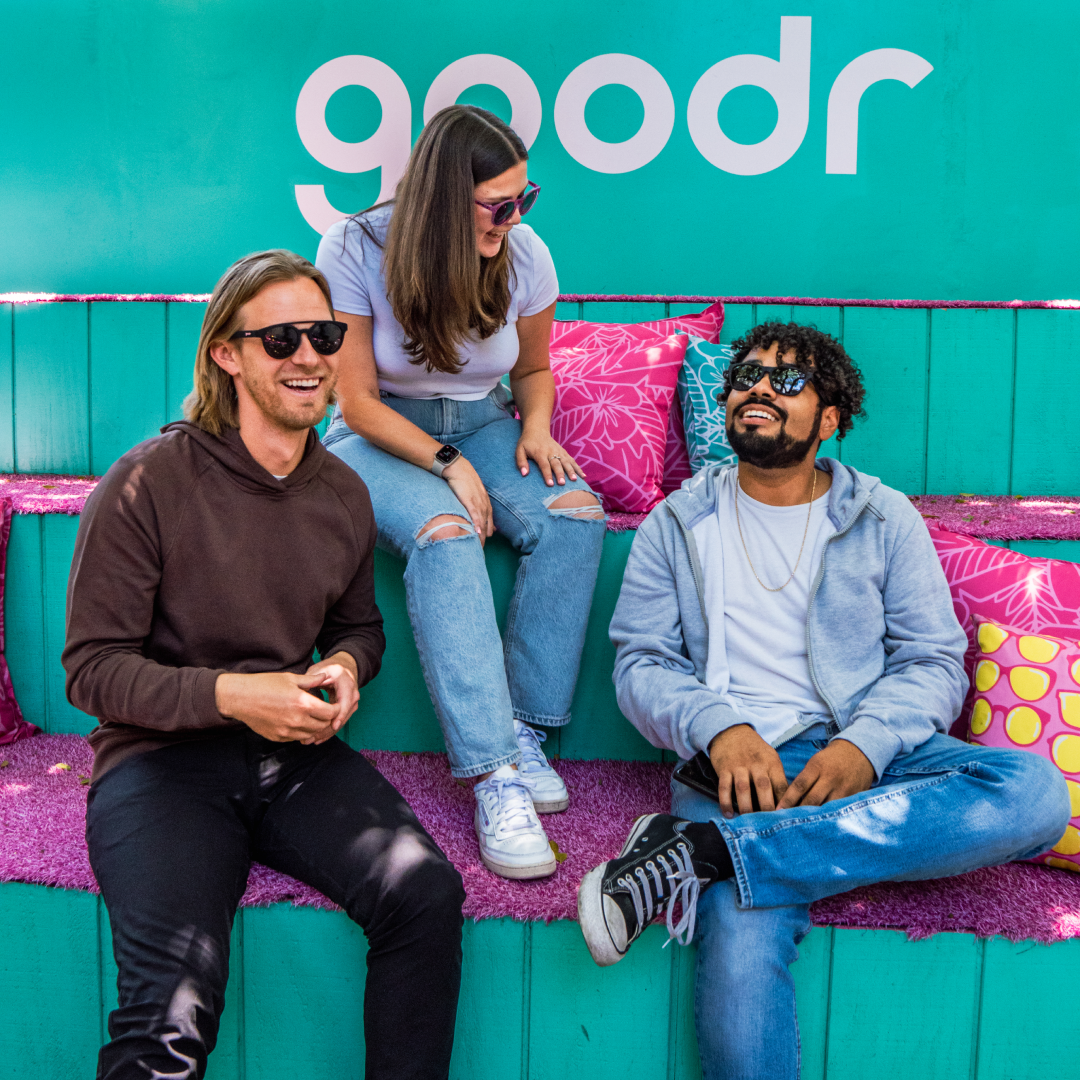 Three friends, all wearing sunglasses, laugh and chat while sitting on colorful steps decorated with vibrant pillows. Behind them is a teal wall with the "goodr" logo. The setting has a relaxed, fun atmosphere.