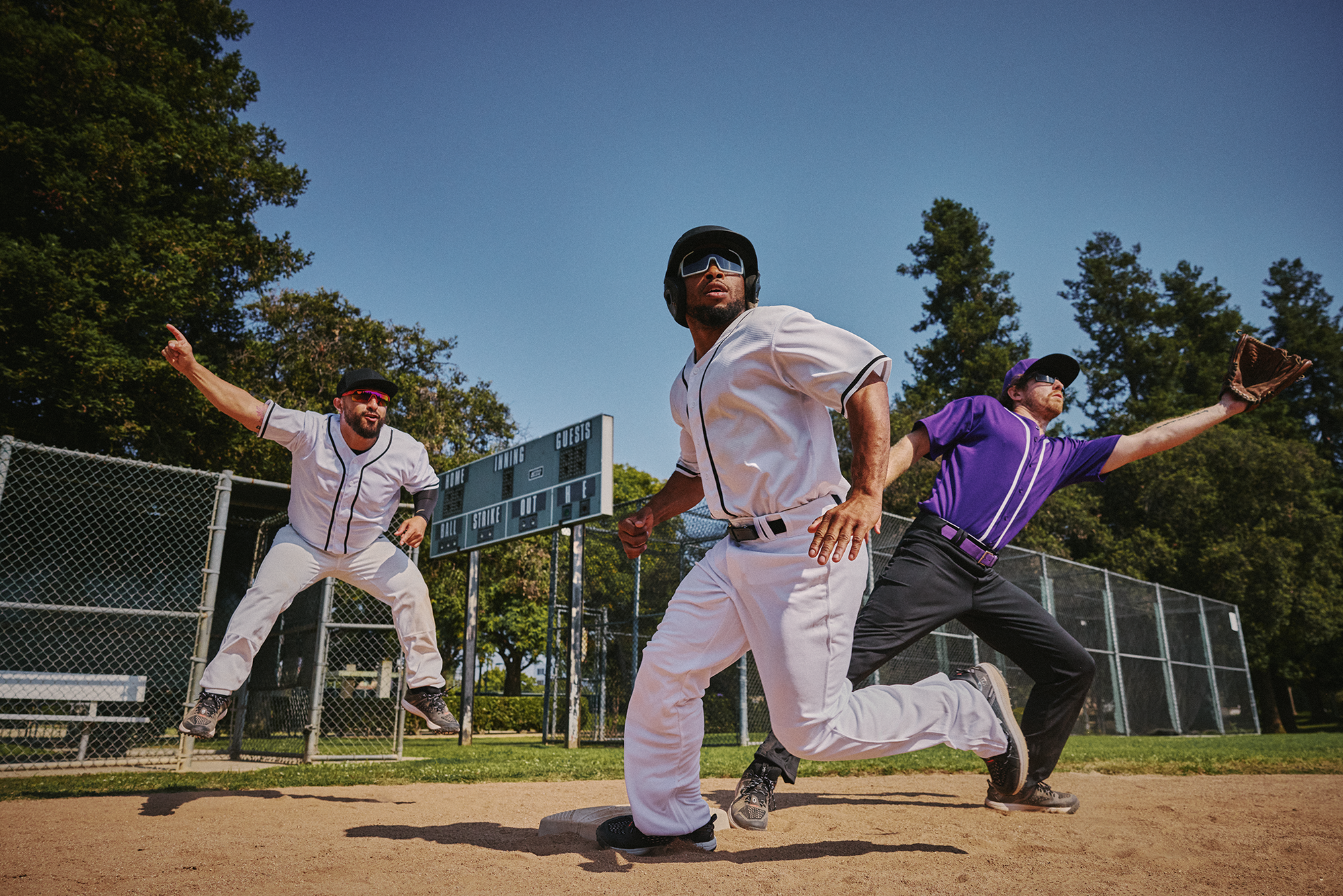 Baseball players wearing sunglasses in mid-action: a runner in white slides towards base as two fielders, one in white and another in purple, attempt to tag or catch the ball on a sunny day at a baseball field.