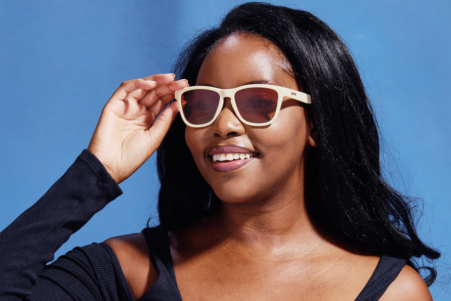 Smiling person wearing beige goodr sunglasses against a blue backdrop.