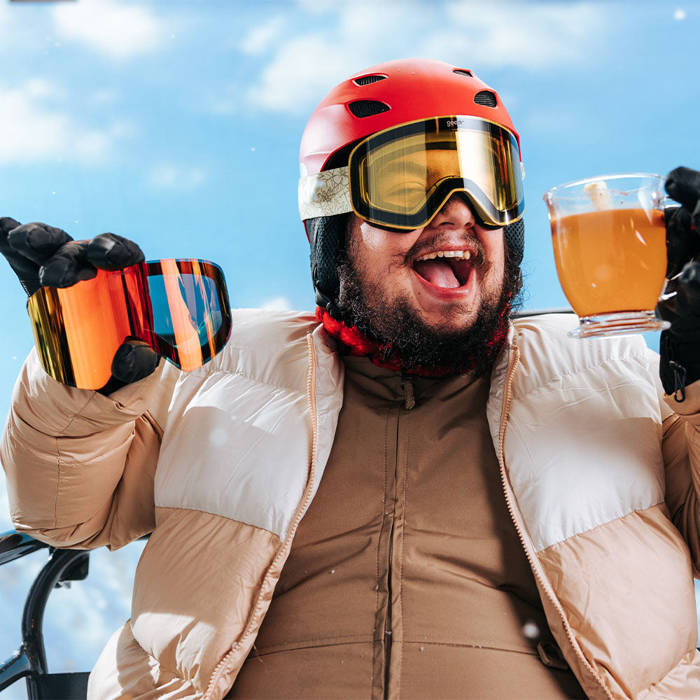 A smiling man wearing a red ski helmet and yellow-tinted ski goggles holds a hot drink in one hand and a pair of reflective orange ski goggles in the other.