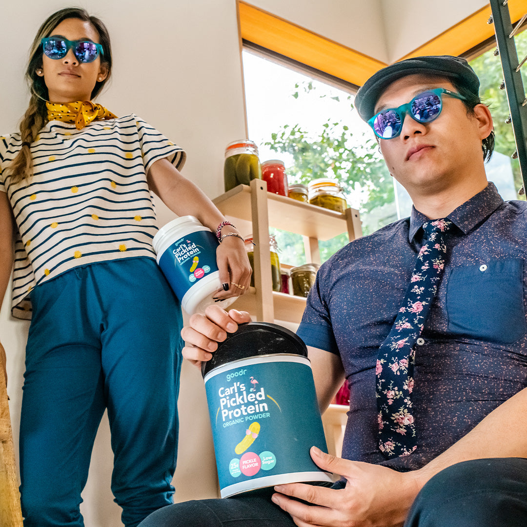 A man and woman wearing blue & purple goodr sunglasses, holding jars of Carl's pickled protein powder