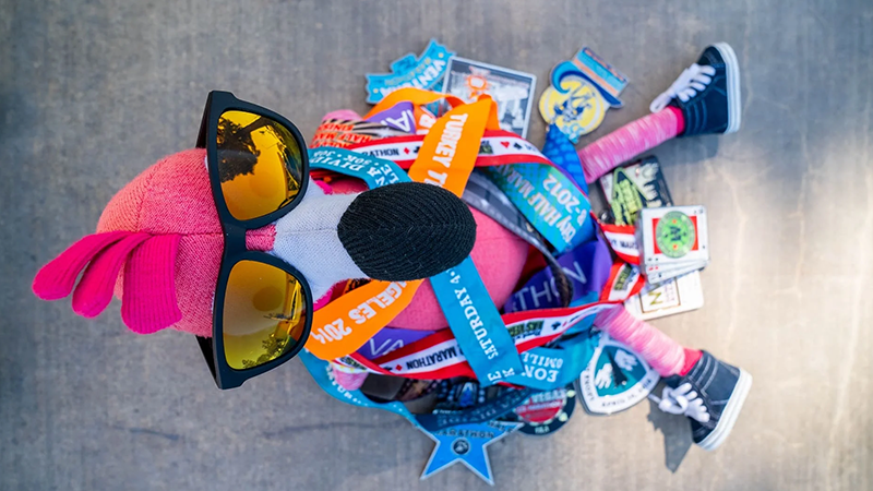 Plush flamingo wearing black sunglasses and surrounded by colorful race medals, with its legs dressed in sneakers, displayed on a concrete surface.