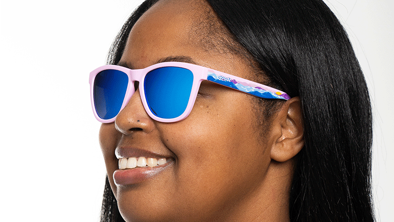 Person smiling while wearing sunglasses with pink frames, blue reflective lenses, and a colorful pattern on the arms.