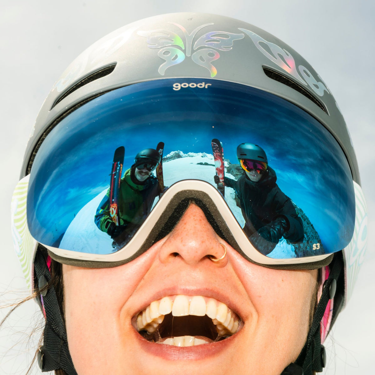 Smiling skier wearing blue mirrored snow goggles and helmet, with two skiers reflected in the lens.