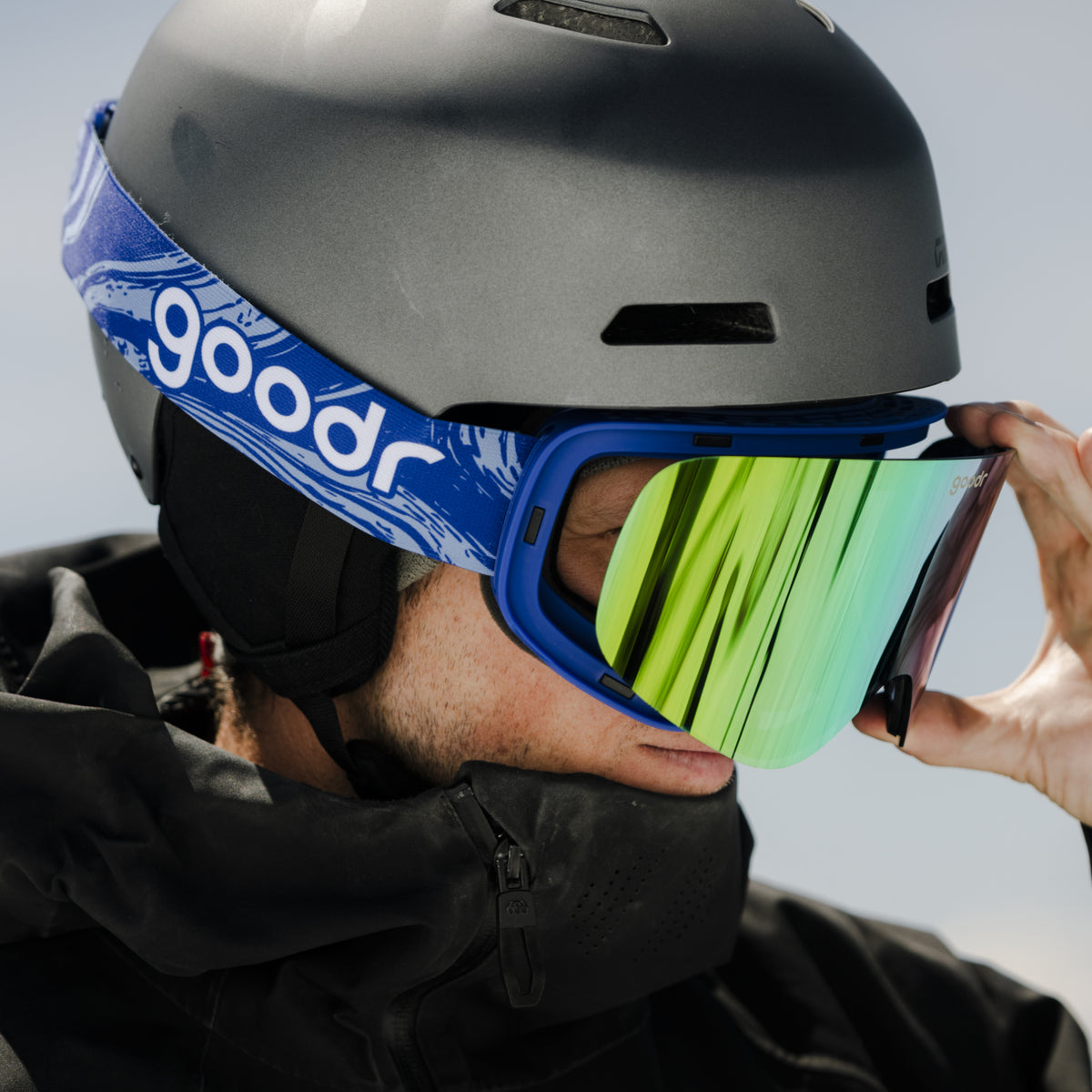 Skier in a black helmet adjusting bright green mirrored goodr ski goggles.
