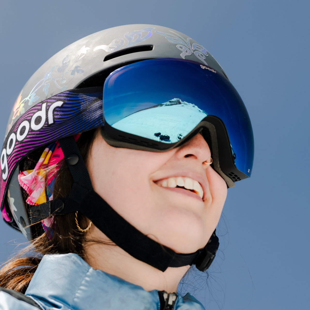 Smiling skier wearing bronze blue mirrored snow goggles with a pink and purple goodr strap and helmet.