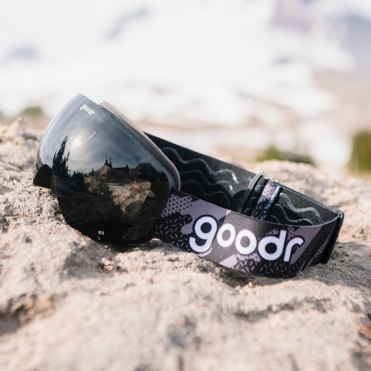 Black snow goggles with a black frame and patterned goodr strap resting on a rock.