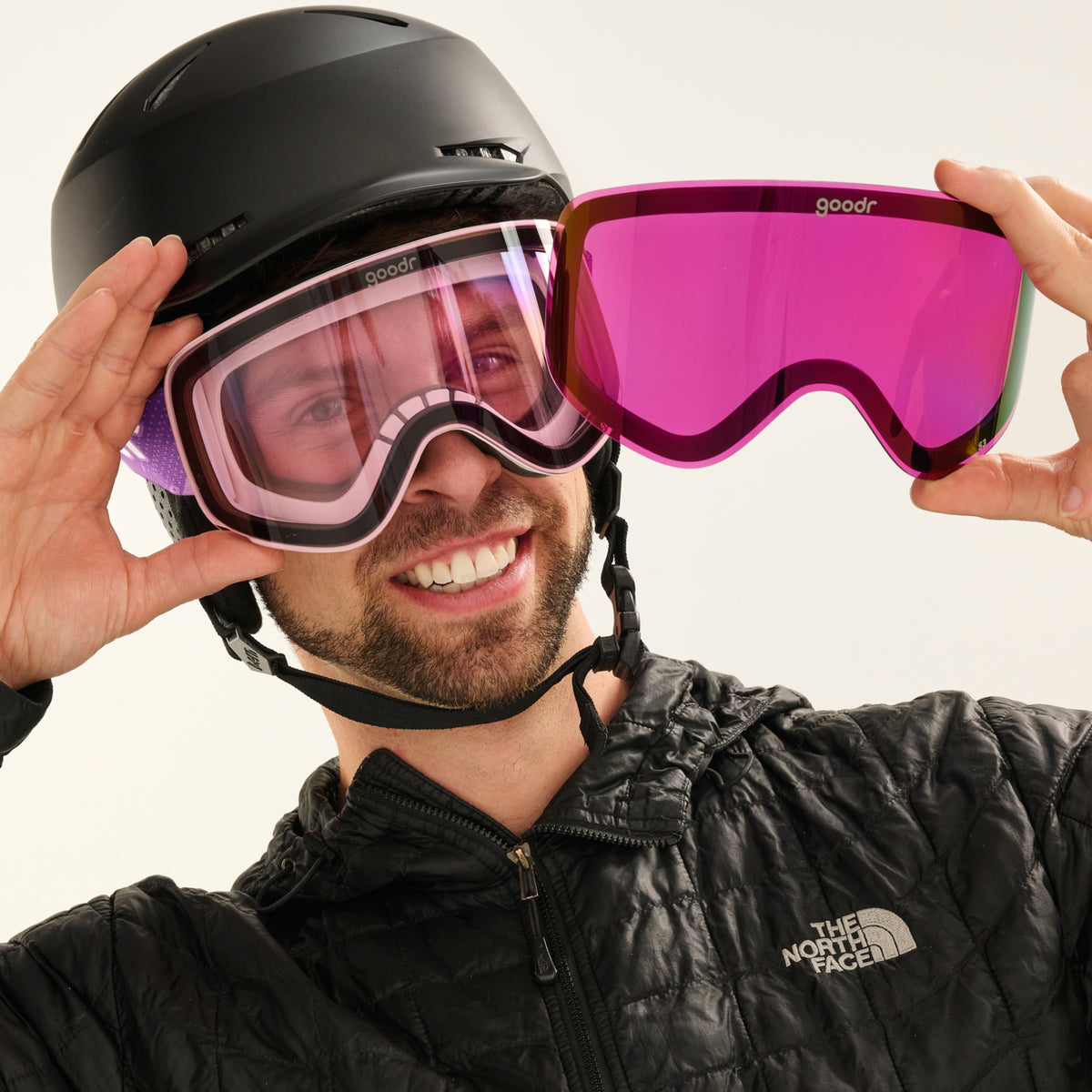 Smiling skier holding black goggles and a pink mirrored goodr replacement lens.