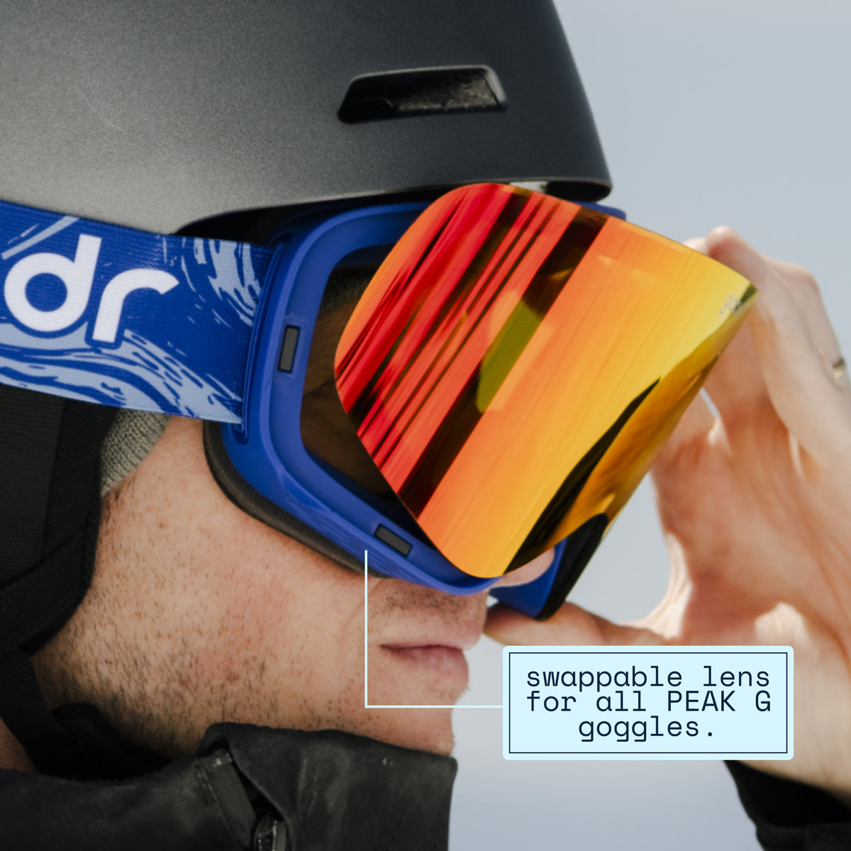 Skier swapping a red-orange mirrored replacement lens onto blue goodr ski goggles.