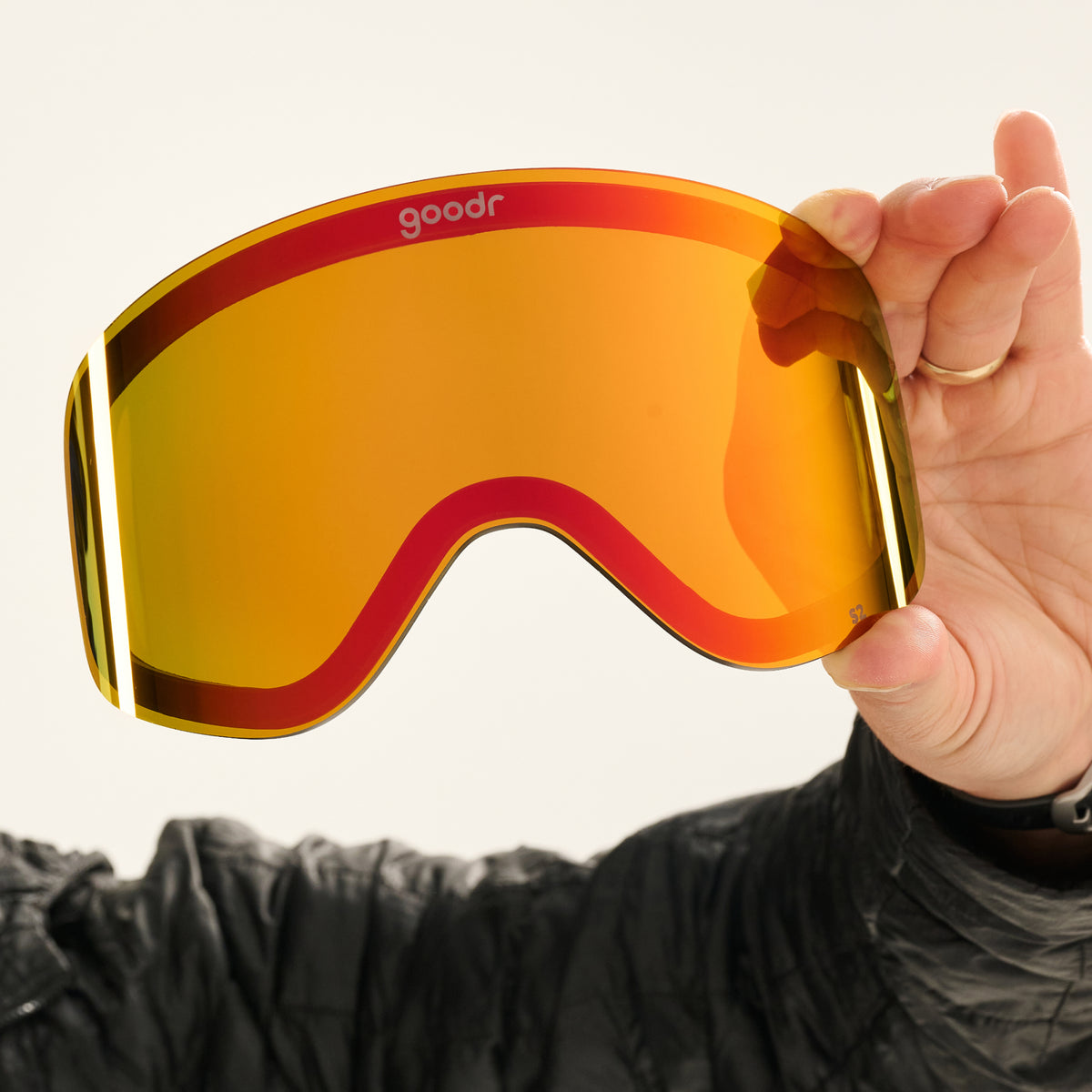 Hand holding a red-orange mirrored goodr replacement lens beside a skier in black.