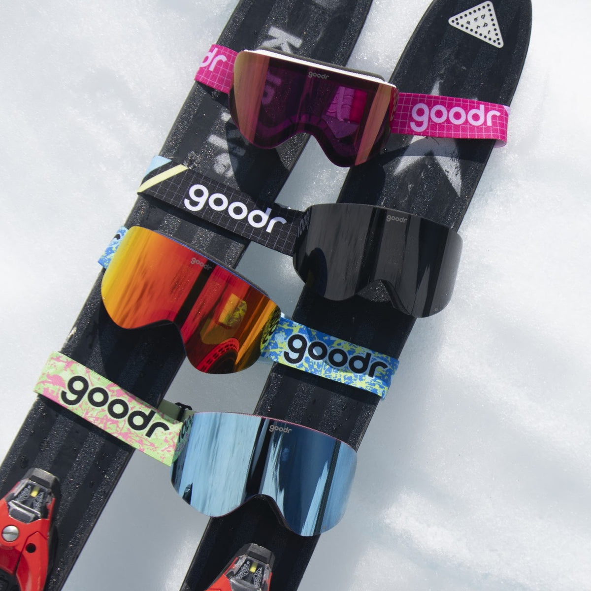 Green and pink goodr goggle strap shown on black ski goggles alongside other colorful goggles on snow.