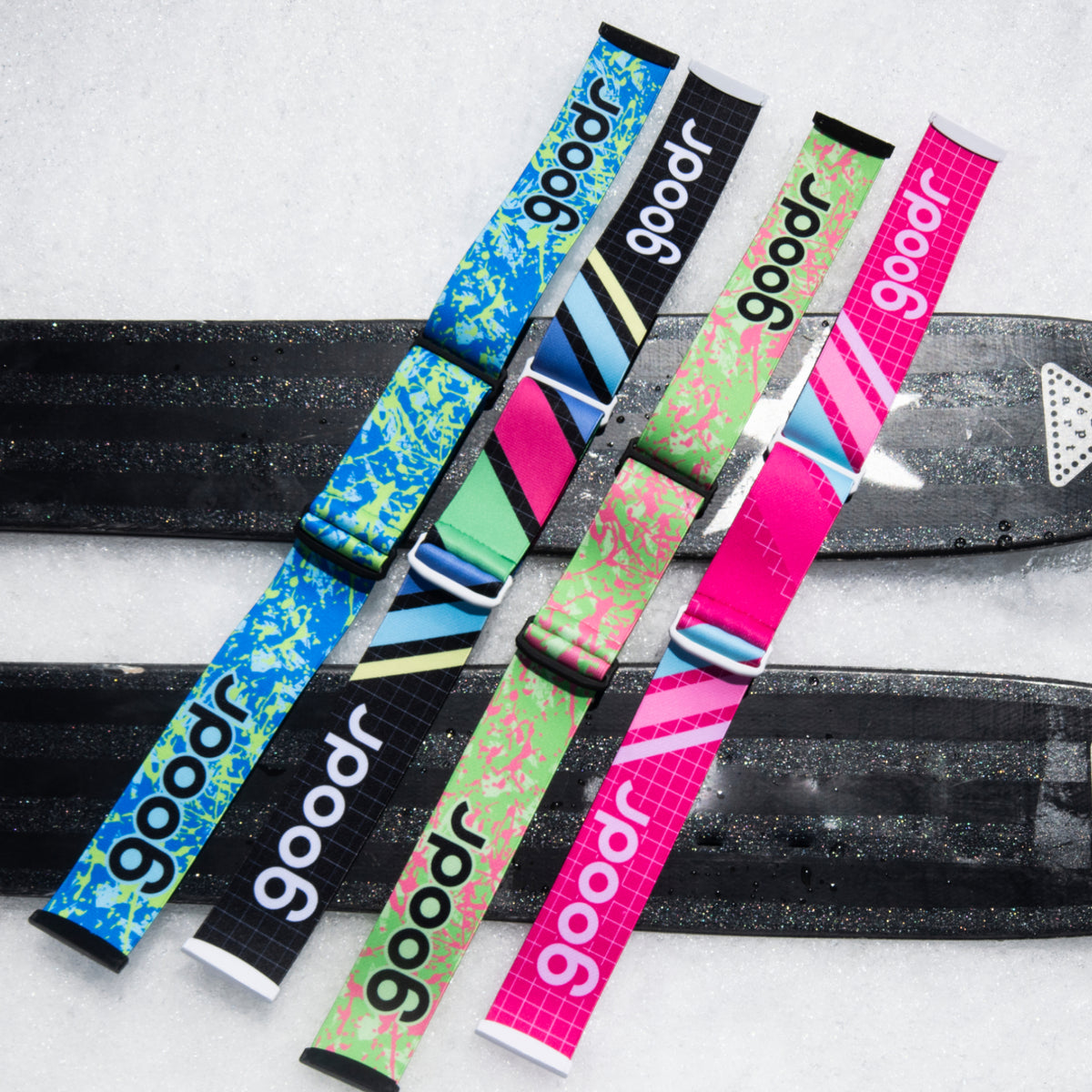 Four colorful goodr replacement goggle straps arranged across black skis in snow.
